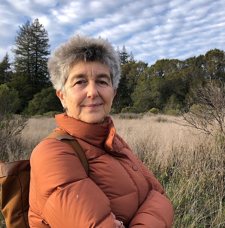 woman in orange-brown jacket standing in field with trees and cloudy sky behind