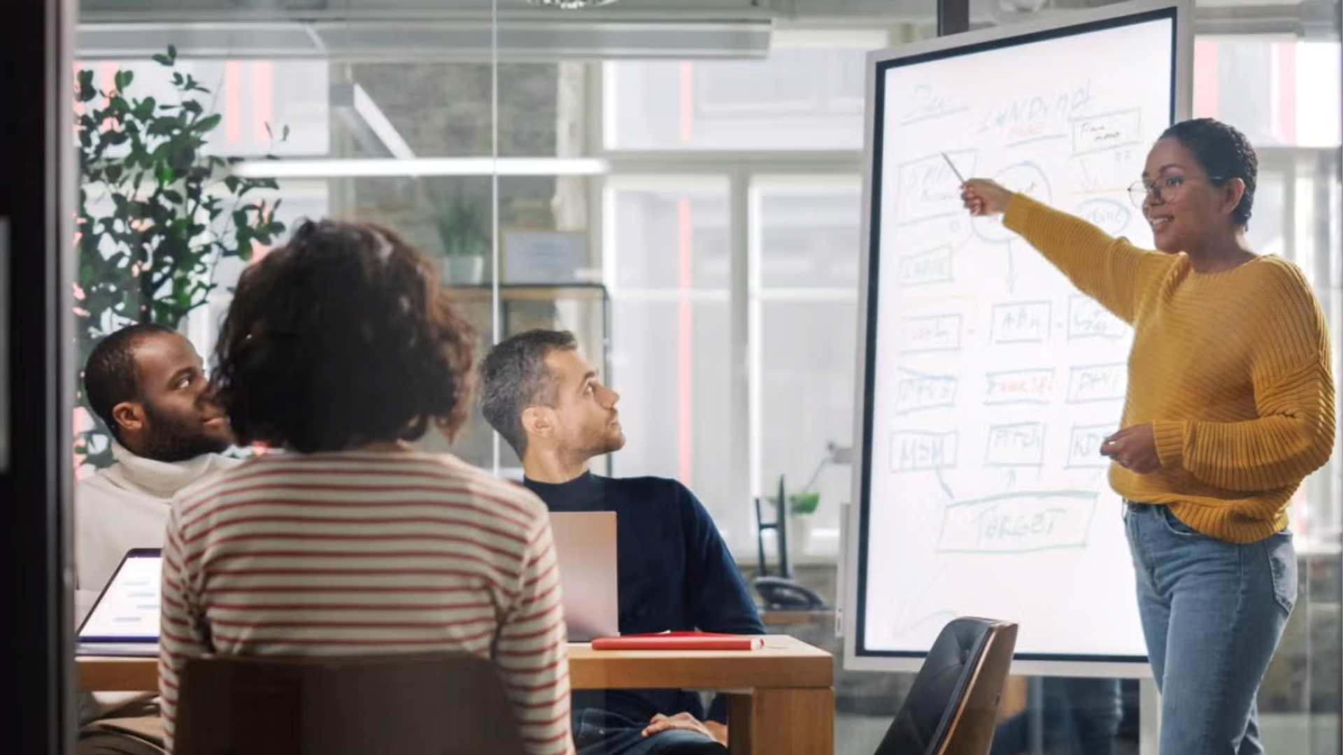 Woman presenting in front of whiteboard in conference room with three people sittig around a conference table.