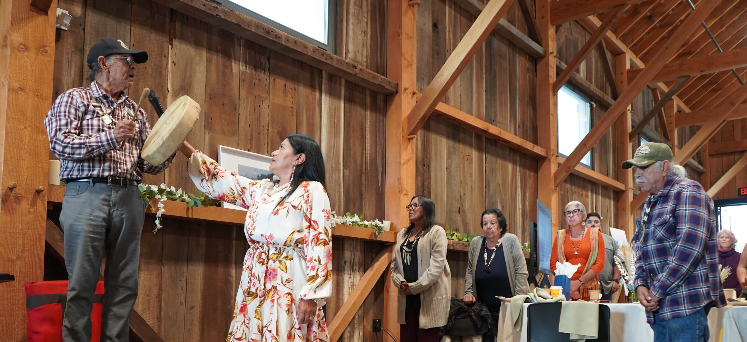 An Indigenous elder holds a traditional drum and speaks into a microphone held by a woman during a cultural presentation at the Cowell Ranch Hay Barn.