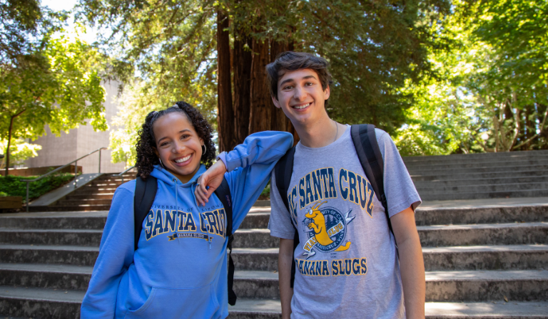 Two students wearing UC Santa Cruz t-shirts smile together with redwood trees in the background