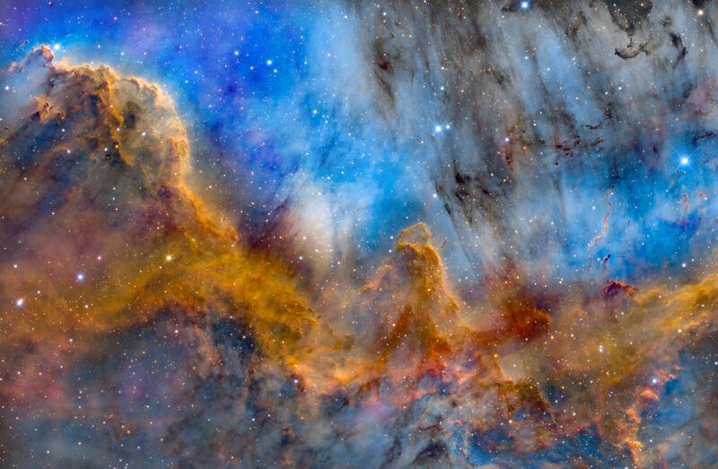 Colorful nebula with swirling clouds of blue, gold, and brown gas and dust, forming pillar-like structures against a dense star field.