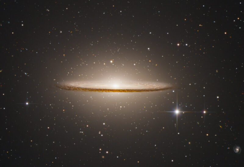 Edge-on spiral galaxy with a bright central bulge and a thin, glowing disk of dust, floating in a dark field filled with distant stars and faint galaxies.