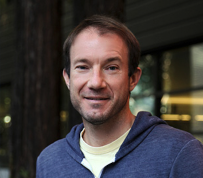 A Caucasian male waring a white shirt and blue jacket stands in front of a tree and a window.