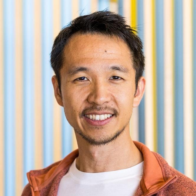 Headshot of Edward Wang. He is an Asian male with a beard, mustache, white shirt, and orange jacket. Behind him is a striped wall of blue, pink, and yellow.
