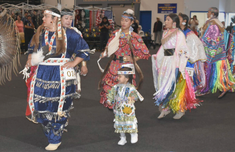A multigenerational group of Indigenous dancers in colorful, traditional regalia participate in a dance at the UCSC Powwow.