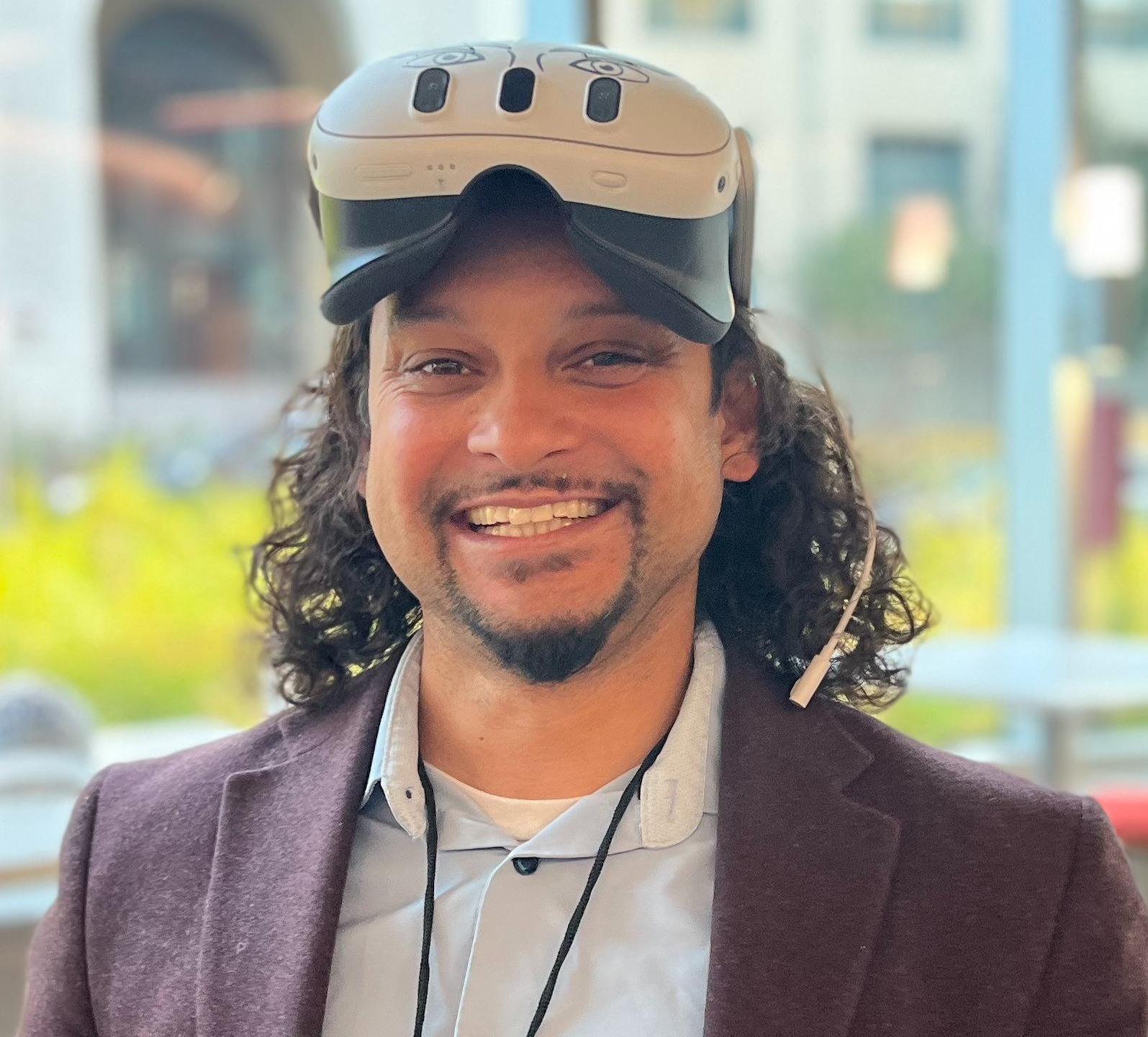 A man wearing a white shirt and purple dress jacket stands in front of a courtyard. He has a VR device on his forehead.