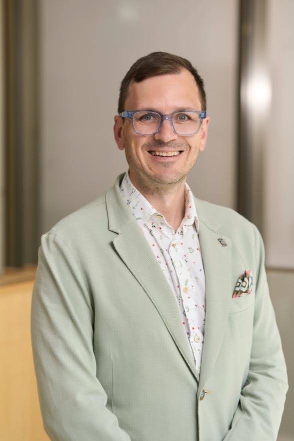 A man with glasses stands in front of a whiteboard. He is wearing a light grey suit jacket and a white button-up shirt.