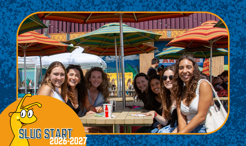 A group of smiling young adults sit gathered around outdoor wooden picnic tables with colorful umbrellas on a sunny day.