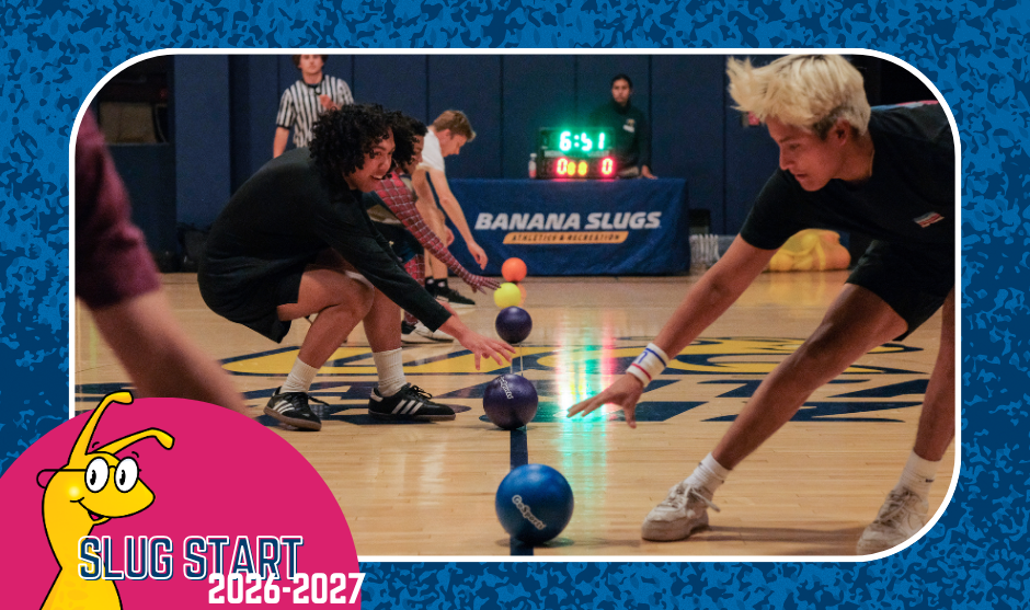 Two players lean forward across the center line to grab dodgeballs lined up on a gymnasium floor during a game in front of a "Banana Slugs" banner.