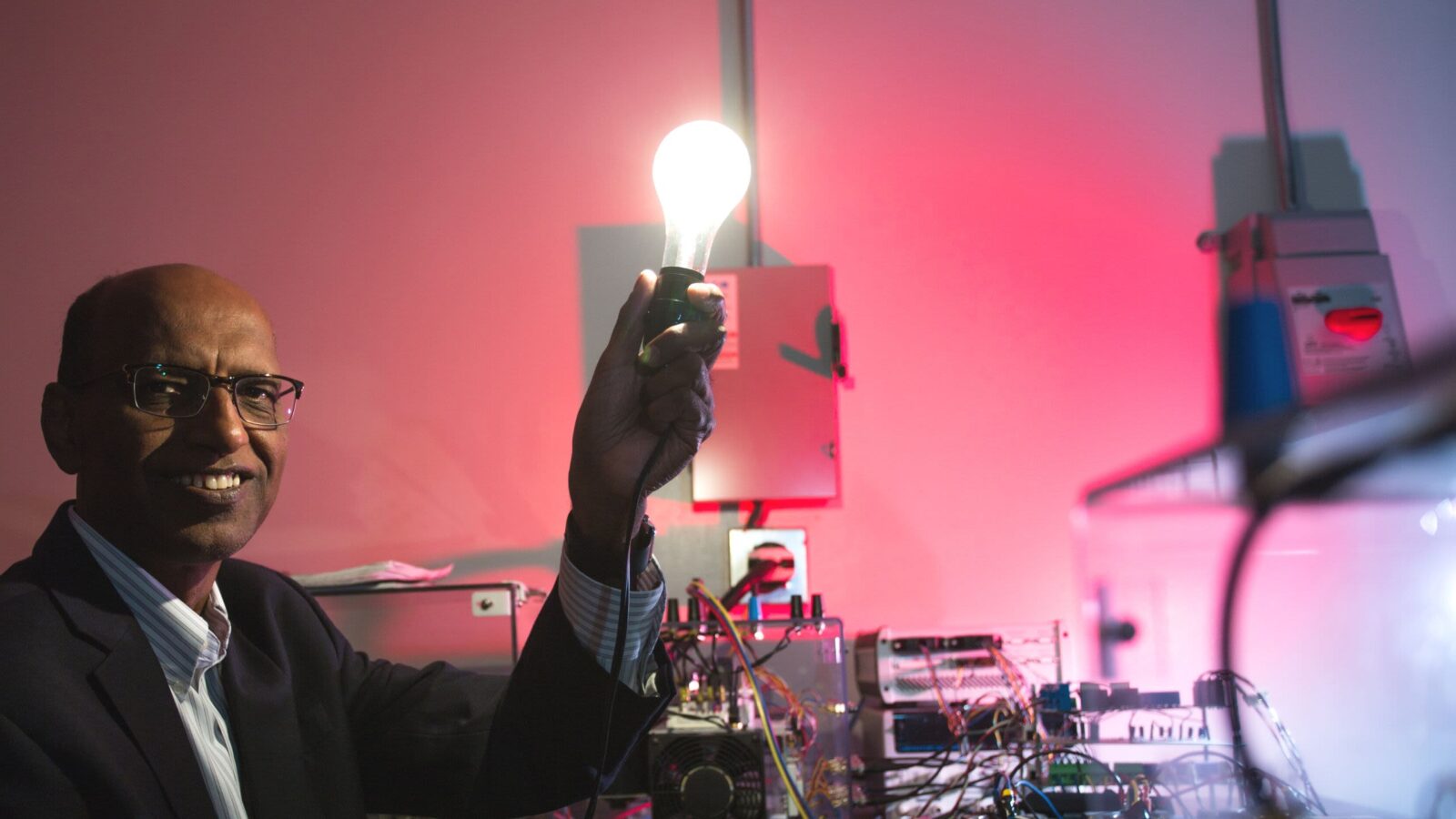 A man in a black suit holds a lightbulb in front of a red background.
