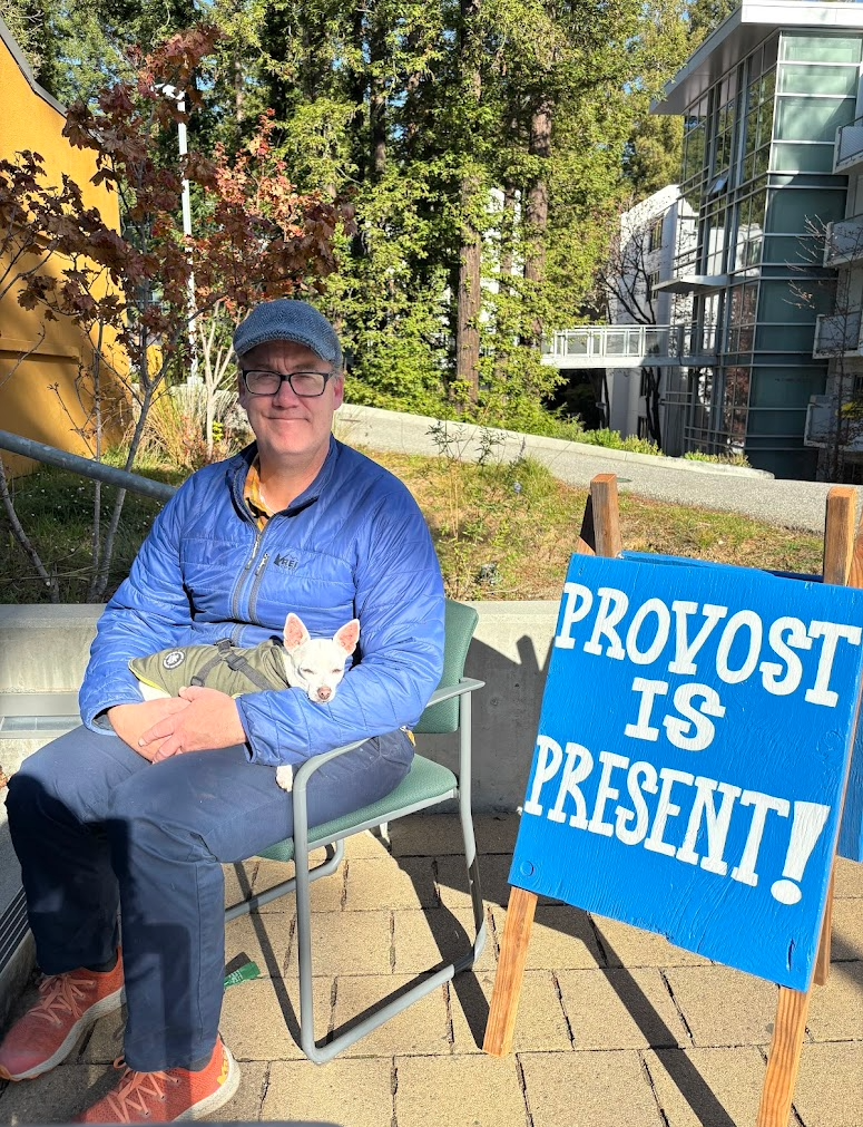 Provost Aims McGuinness holds Poppy the Chihuahua while seated by the Provost is Present sign.