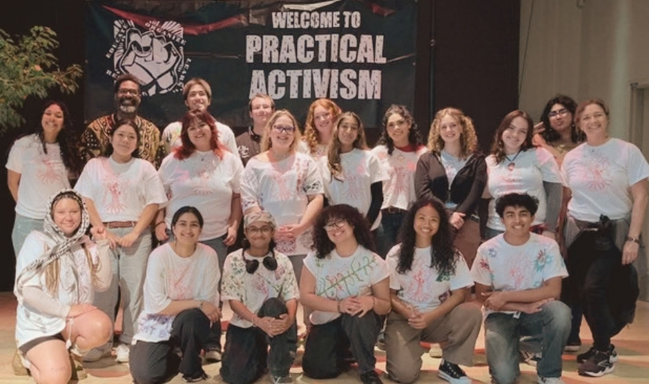 A large, diverse group of smiling students poses together in front of a banner reading "Welcome to Practical Activism," with many wearing matching white t-shirts.